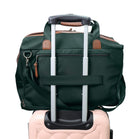 A dark green rolling duffel bag with brown leather accents and an extended silver telescoping handle, shown resting on top of a light pink luggage.