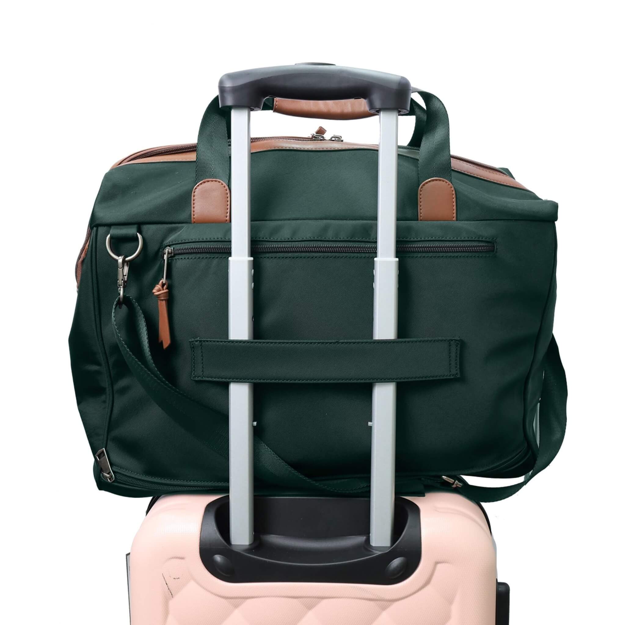 A dark green rolling duffel bag with brown leather accents and an extended silver telescoping handle, shown resting on top of a light pink luggage.